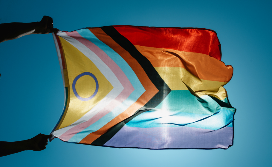 Two hands emerging from the left hold a pride progress flag against the sun, which shines through the sheer rainbow-coloured material against a clear blue sky.