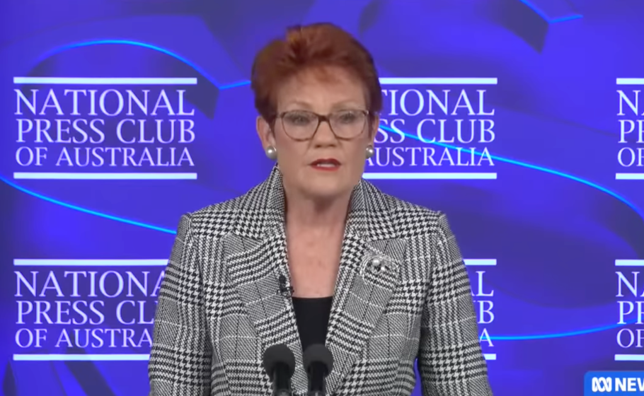 A woman with short red hair and glasses, wearing a black and white houndstooth jacket, stands at a podium with microphones in front of a blue backdrop with the words "National Press Club of Australia" repeated.